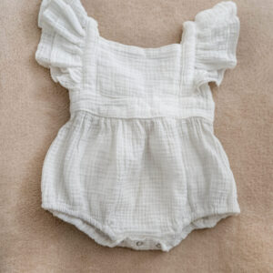 White muslin baby romper with flutter sleeves and bow tie back on soft background.