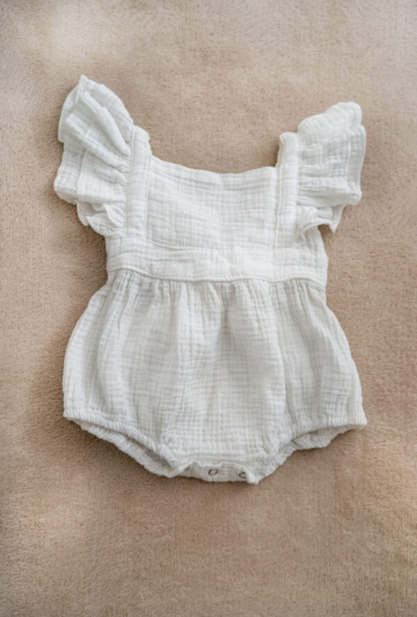 White muslin baby romper with flutter sleeves and bow tie back on soft background.