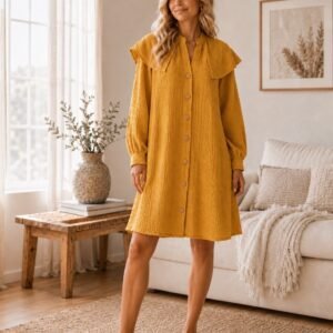 Woman wearing a mustard cotton muslin shirt dress with ruffle collar, long sleeves and button front